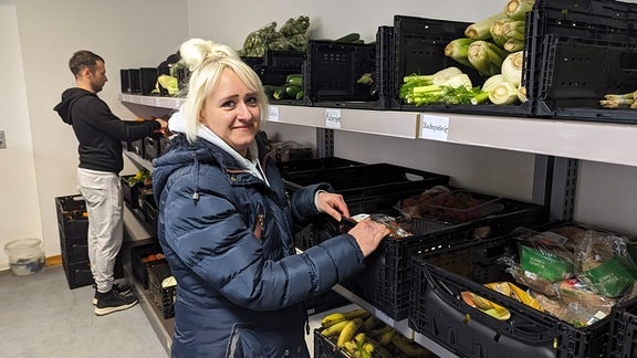 Die Tafel-Mitarbeiterin Jennifer Siermann steht vor einem großen Lebensmittelregal in der Tafel Weißenfels. Im Hintergrund ein Mitarbeiter.