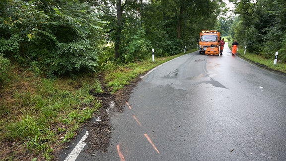 Eine Straße mit Kennzeichnung des Unfallhergangs und einem orangefarbenen Spezialfahrzeug im Hintergrund