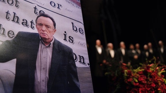 Ein Bild des Schauspielers Peter Sodann ist an einem Rednerpult im Neuen Theater aufgestellt, während ein Chor singt.