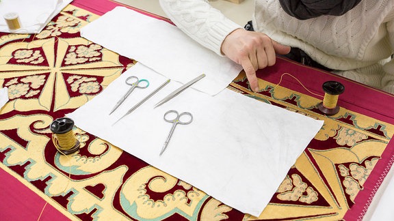 Eine Hand auf einem Textil, dazu Scheren, Pinzetten und Garne.