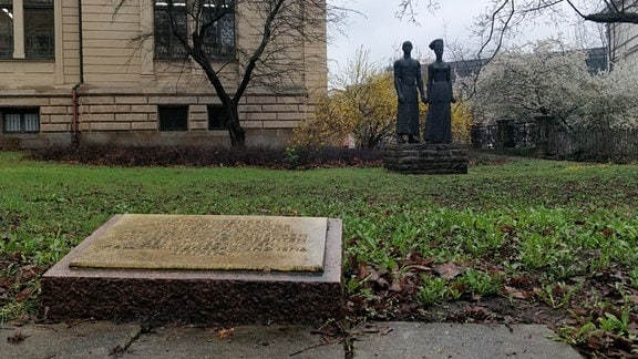 Im Vordergrund eine Gedenktafel auf dem Boden, rundherum eine Wiese, im Hintergrund zwei Statuen und ein Gebäude