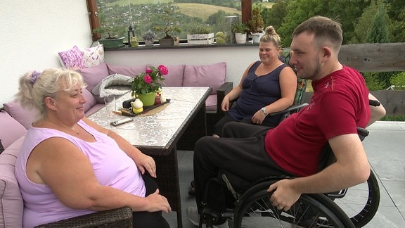 Zwei Frauen sitzen zusammen mit einem Mannin Rollstuhl auf einer Terasse.