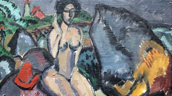 Ein Gemälde von Ernst Ludwig Kirchner: Eine nackte Frau ist zwischen grauen und bunten geometrischen Formen zu sehen, die wie Felsen anmuten. Im Hintergrund ist eine Landschaft mit Bäumen und Gebäuden angedeutet.