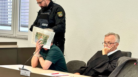 Der Angeklagte (M) sitzt im Gerichtssaal neben seinem Anwalt Wolfgang Müller (r) und verdeckt sein Gesicht.