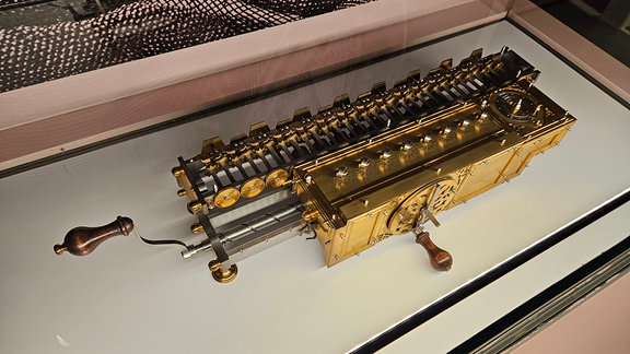 Eine goldene Maschine steht in einer Glasvitrine. Eine Kurbel bringt den komplizierten Mechanismus in Gang. Dies ist eine Rechenmaschine aus dem 17. Jahrhuntert. 
