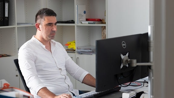 Ein Mann mit dunklen Haaren und weißem Hemd sitzt vor einem Computer an einem Schreibtisch