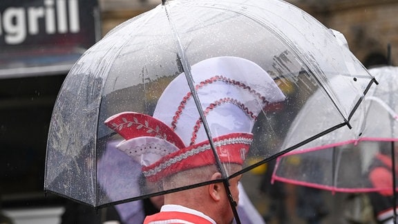 Ein Karnevalist mit Karnevalskappe steht unter einem Regenschirm. Es regnete zur Eröffnung der Karnevalssession dauerhaft. Traditionell zum 11.11. übernehmen die Narren der Saalestadt das Rathaus. Diesmal musste das Programm durch den anhaltenden Regen geändert werden.