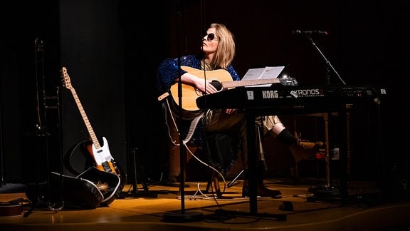 Multi-Instrumentalistin Wencke Wollny, eine Frau mit langem, dunkelblondem Haar, Sonnenbrille und blauer Glitzerjacke, sitzt hinter einem Keyboard und hält eine Gitarre in der Hand.