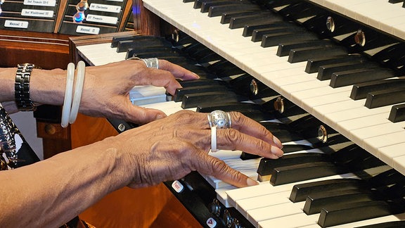 Zwei mit Ringen und Armbändern verzierte Hände spielen auf einer klavierähnlichen Tastatur mit weißen und schwarzen Tasten   