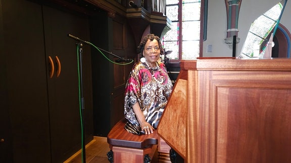 Eine ältere Frau in einem bunt gemusterten Oberteil sitzt an einer Orgel und lächelt in die Kamera 