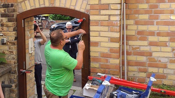 Ein Mann schraubt an einem Rahmen für eine neue Tür an der Synagoge in Halle, zwei Männer stehen daneben.