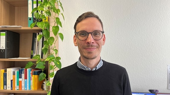 Oliver Winkler, ein Mann mit Brille und schwarzem Pullover, steht in einem Büro vor einem Bücherregal und lächelt in die Kamera