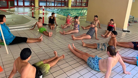 Kinder sitzen auf dem Boden in der Schwimmhalle und üben Beinbewegungen