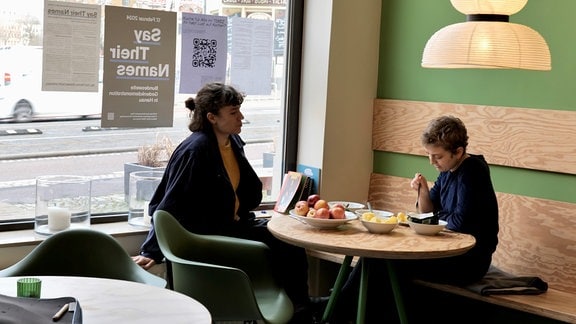 Zwei Personen sitzen in einem Café an der Fensterscheibe, auf dem Tisch steht Essen.