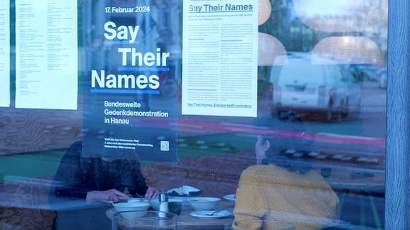 In einem Schaufenster hängen Plakate, dahinter sitzen zwei Personen.