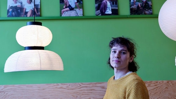 Eine Frau sitzt auf einer Bank vor einer grünen Wand und blickt direkt in die Kamera.