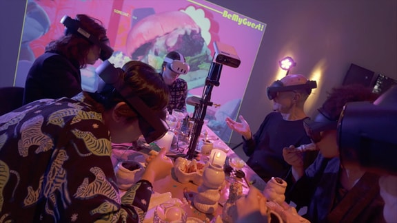 Mehrere Menschen sitzen mit VR-Brillen an einem Esstisch, an der Wand eine Projektion, auf der ein Burger zu sehen ist und der Schriftzug "Be My Guest" (auf Deutsch: Sei mein Gast)