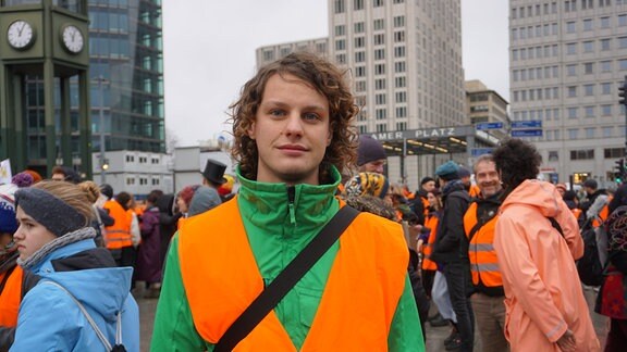 Ein junger Mann steht auf einer Demonstration. Er trägt eine orangefarbene Weste und blickt direkt in die Kamera.