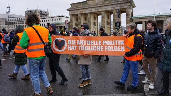 Eine Gruppe der Letzten Generation vor dem Brandenburger Tor in Berlin. Die Menschen in orangefarbenen Warnwesten tragen ein Transparent mit der Aufschrift "Die aktuelle Politik hat tödliche Folgen".