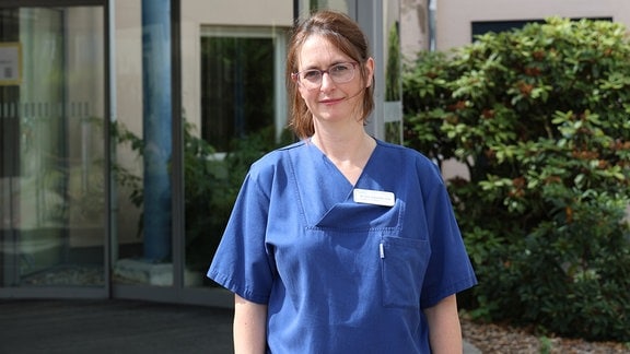 Eine Ärztin steht in blauem Kittel vor der Eingangstür eines Krankenhauses in Sangerhausen.