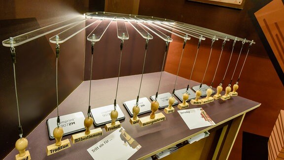 In einer Ausstellung hängen verschiedene Stempel; darunter ist ein Tisch mit Stempelkissen und Papier zu sehen. 