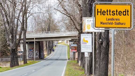 Neben einer leeren Straße stehen mehrere Bäume, außerdem ein Ortsschild von Hettstedt im Landkreis Mansfeld-Südharz