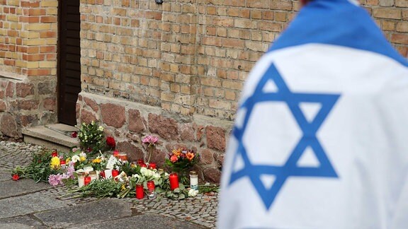 Ein Mann mit einer Israel-Flagge steht vor der Synagoge, wo Blumen und Kerzen stehen und liegen