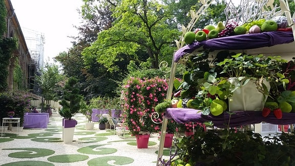 Blick in den Paradiesgarten der Neuen Residenz mit rosa Pflanzen, fliederfarbenen Tischdecken. Im Vordergrund steht eine große Etagere mit viel Obst. 