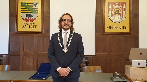 Der neue Merseburger Oberbürgermeister Sebastian Müller-Bahr steht mit einer Kette um den Hals zwischen zwei Wappen in einem Rathauszimmer.