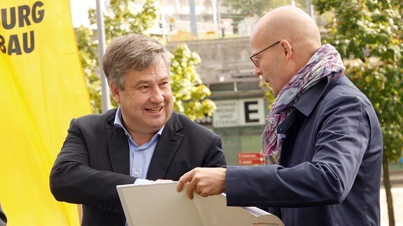 Ein Mann überreicht einem anderen Mann eine weiße Mappe mit Dokumenten. 