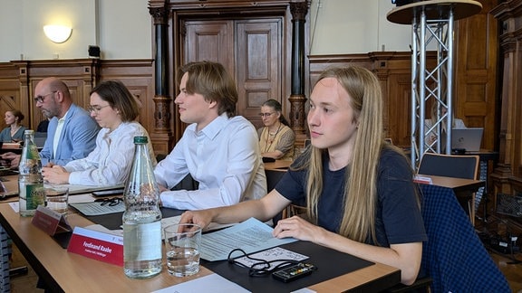 Ein junger Mann mit langen Haaren sitzt in einem Parlament. 