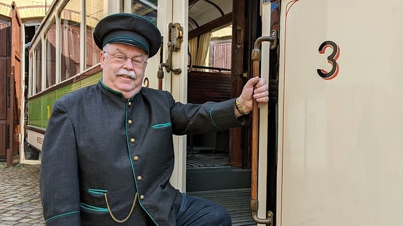 Ein Mann in einer historischen Straßenbahnfahrer-Uniform steht an der Fahrertür eines Triebwagens. 