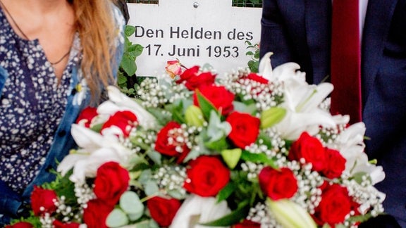 Nahaufnahme zweier Personen, die einen Blumenstrauß halten. Im Hintergrund ein Kreuz mit der Aufschrift "Den Helden des 17. Juni 1953".
