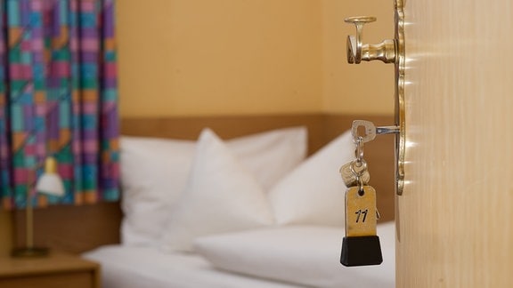 Ein Zimmerschlüssel hängt vor einem Hotelzimmerbett im Türschloss. 