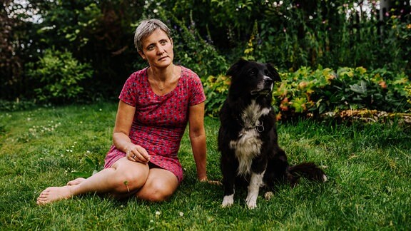 Eine Frau in einem roten Kleid sitzt auf einer Wiese in einem Garten. Neben ihr sitzt ein schwarzer Hund.