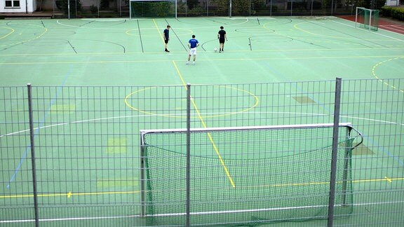 Sportplatz einer Schule in Zeiten von Corona, mit drei Jungen, die Fussball spielen.