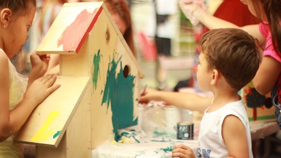 Zwei Mädchen und ein Junge malen im Kindergarten ein Vogelhaus mit grüner Farbe an.