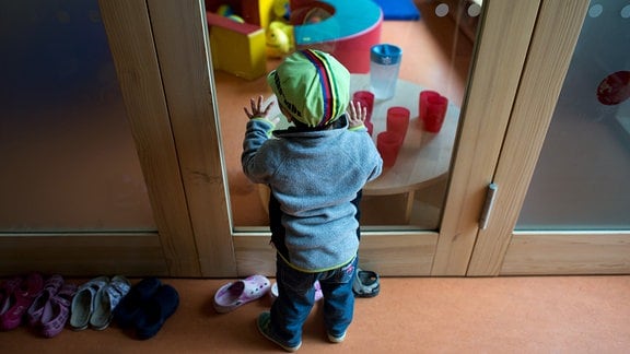 Ein Kleinkind steht 2015 an einer Glasscheibe in einer Kindertagesstätte und schaut anderen Kindern beim Spielen zu.