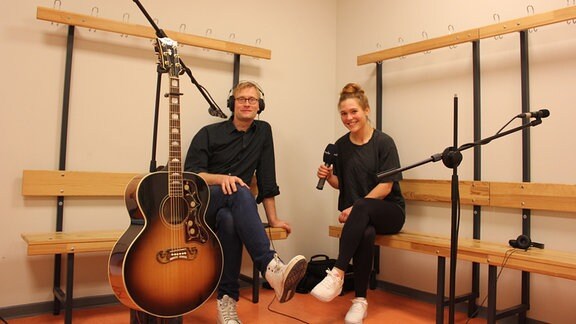 Ein Mann und eine Frau sitzen mit Mikro und Gitarre in einer Umkleidekabine.