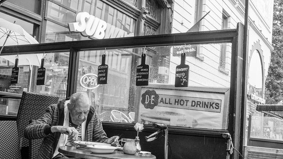 Streetfotografie: Kunst im Funkhaus "Life is live" von Fotograf Wenzel Oschington (Matthias Pavel) – auf dem Bild: Abendessen, älterer Mann (mit Rollator ) im Bistro in Rotterdam 2017