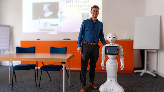 In einem Seminarraum steht ein junger Mann neben einem humanoiden Roboter