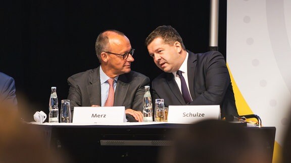 Friedrich Merz und Sven Schulze sitzen nebeneinander an einem Tisch und sprechen miteinander