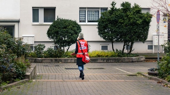 Eine Frau mit roter Umhängetasche und roter Warnweste mit der Aufschrift "Hier ist Die Linke" geht zu einem Wohnblock
