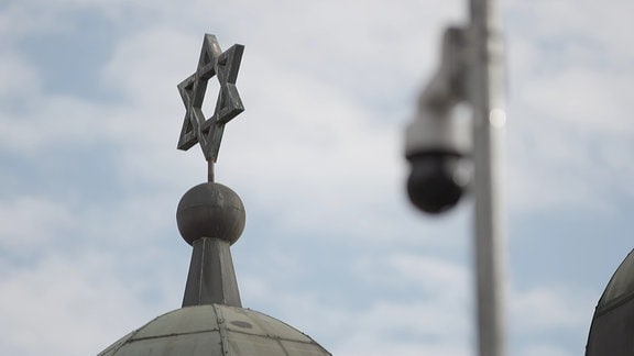 Ein Davidstern auf einem Kuppeldach und blauem Himmel, verschwommen davor auf gleicher Höhe eine kugelförmige Überwachungskamera. 
