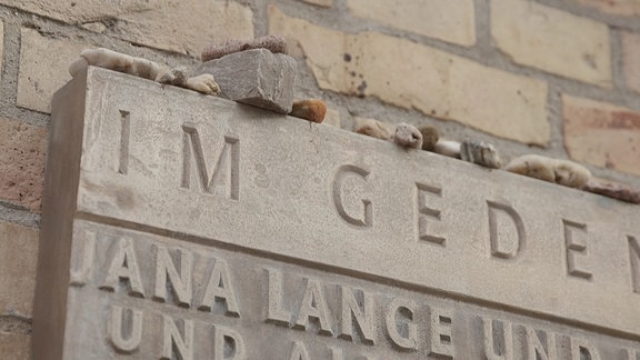 Nahaufnahme der steinernden Gedenktafel an der Synagogenmauer auf der gestapelt Steine liegen. 