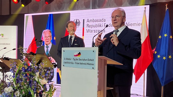 Ministerpräsident Reiner Haseloff steht auf einer Bühne und hält eine Rede. Die Bühne ist mit Blumen und den Flagen der EU, Deutschland und Polen geschmückt. 