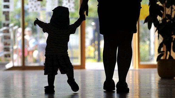 Ein kleines Kind wird in einem Kindergarten von einer Kindergärtnerin an der Hand geführt. 