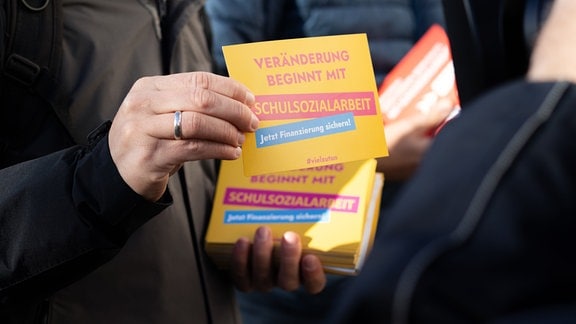 Eine gelbe Postkarte mit der Aufschrift "Veränderung beginnt mit Schulsozialarbeit - Jetzt Finanzierung sichern!" wird in die Kamera gehalten.