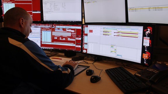 Ein Mann mit Trainingsjacke sitzt vor sechs verschieden großen Bildschirmen, hat zwei Mäuse und zwei Tastaturen vor sich