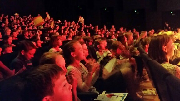 In einem in rotes Licht getauchten Theatersaal sitzen zahlreiche Kinder, die während einer Aufführung klatschen